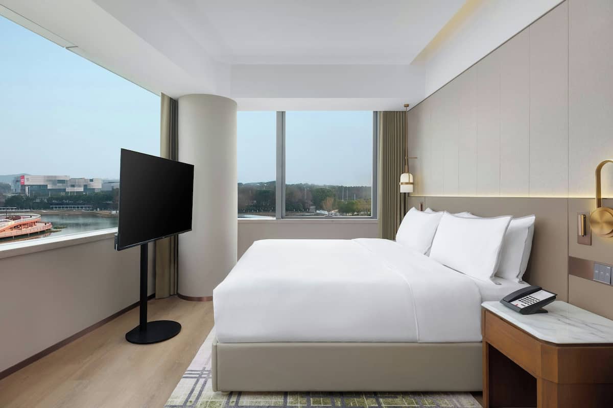 Habitación de hotel con una cama grande, un televisor sobre un mueble, una mesita de noche con un teléfono, y vistas a un cuerpo de agua y edificios.