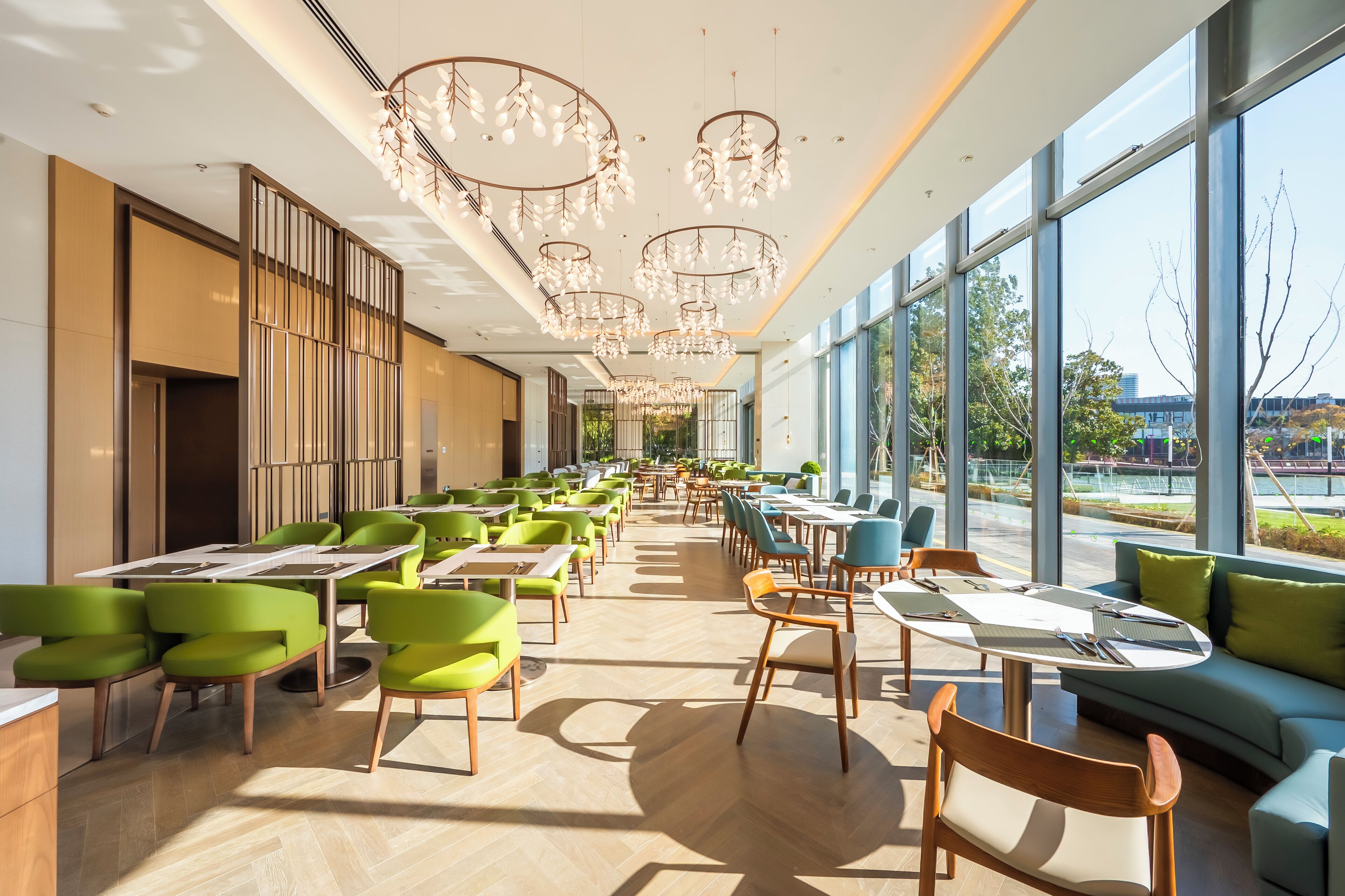 A modern dining area with green chairs, white tables, and large windows offering views of the outdoors.
