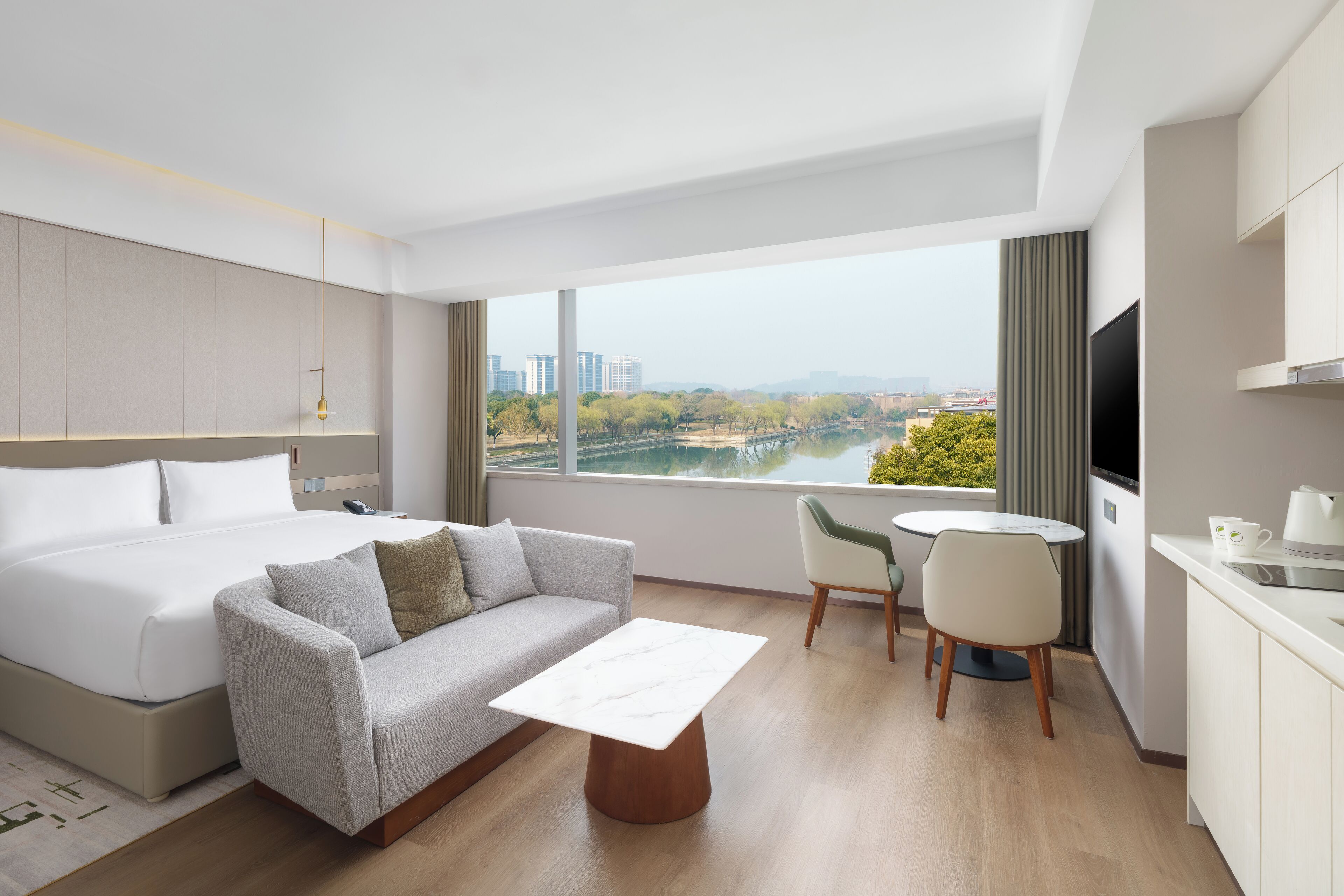 A modern hotel room with a bed, sofa, dining table, chairs, and a TV.