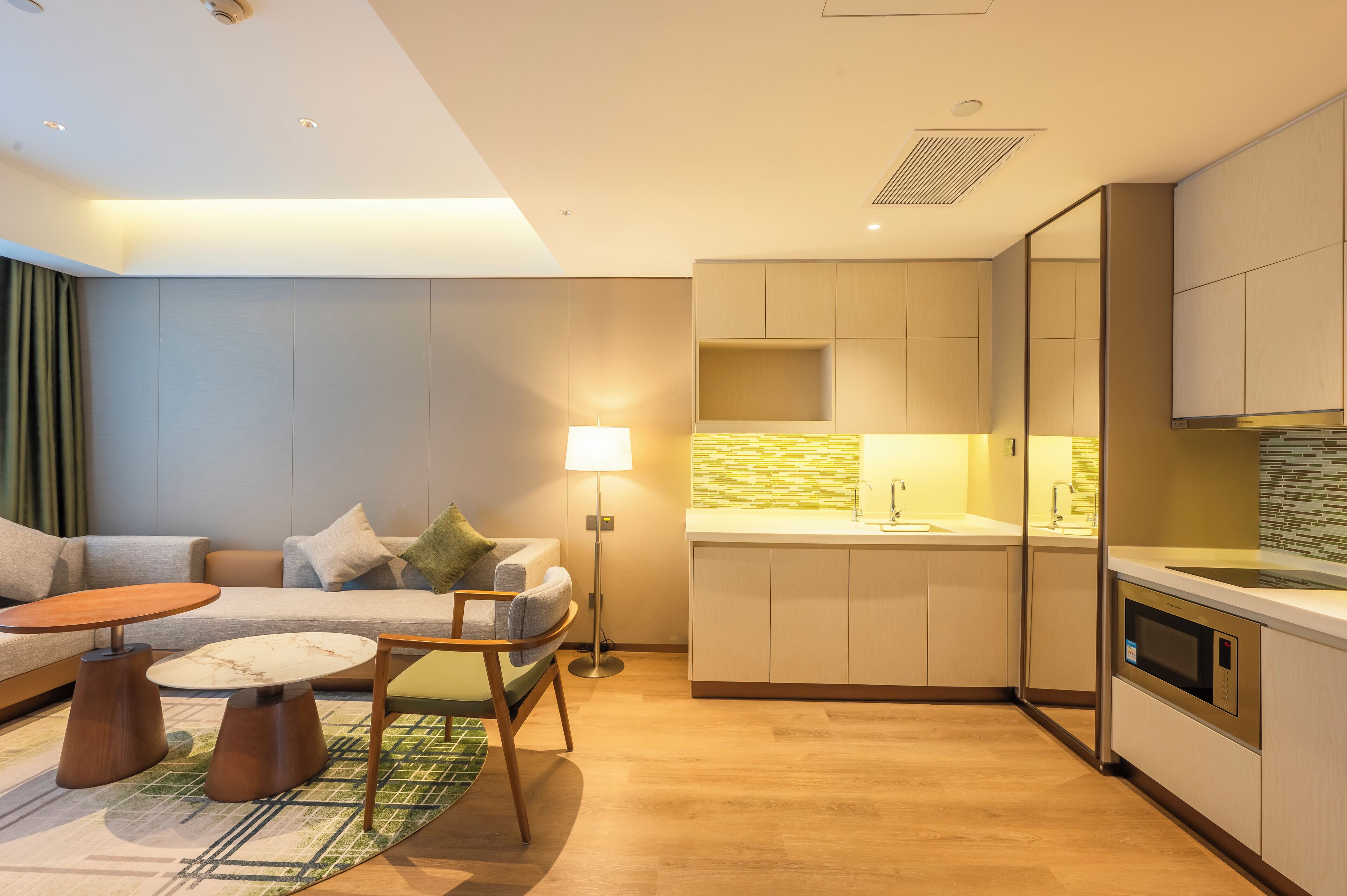 A modern living room with a sofa, coffee table, and lamp, adjacent to a kitchen with a microwave and cabinets.
