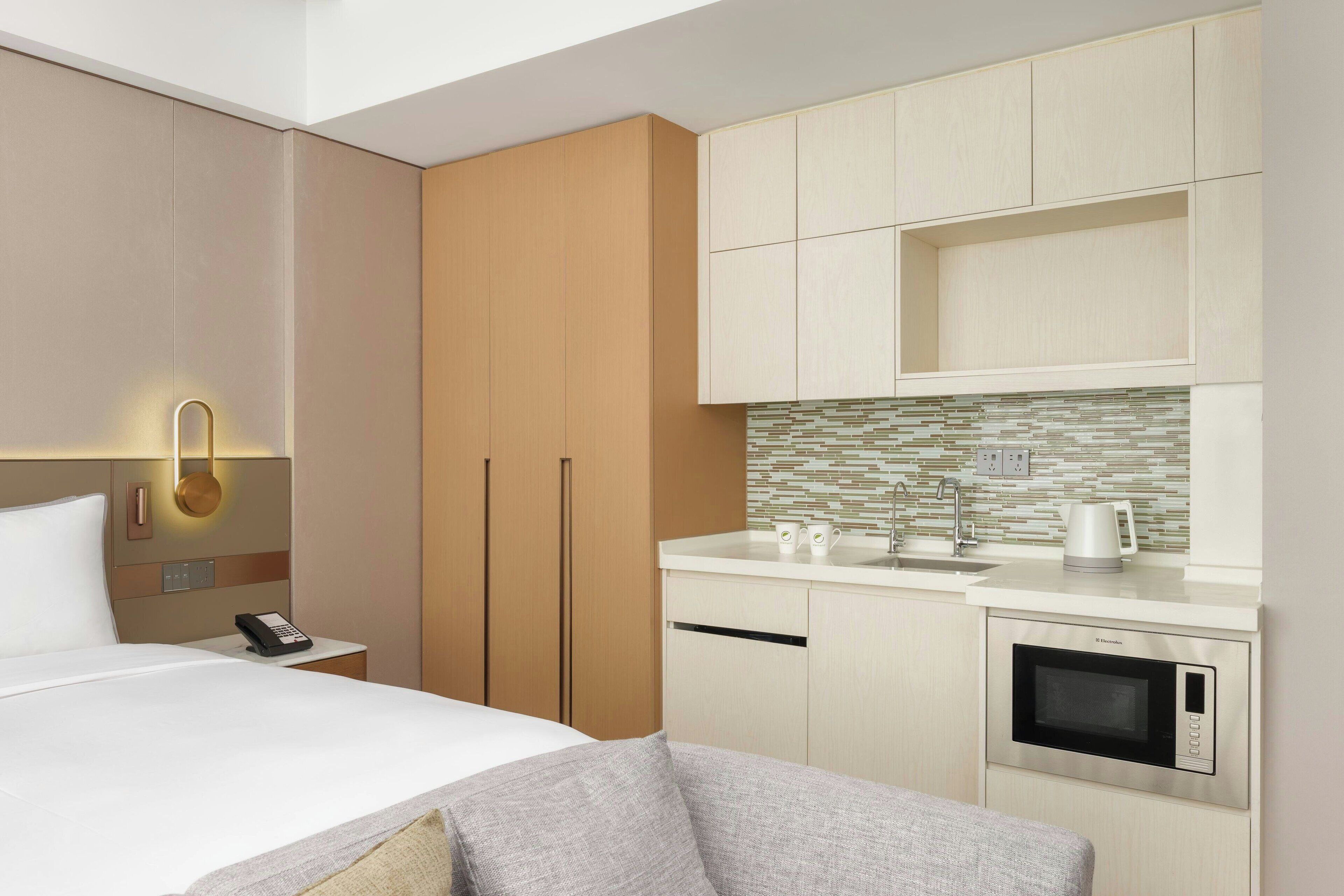 A modern hotel room with a bed, bedside table with a phone, a sofa, a kitchenette with a microwave, and a tiled backsplash.