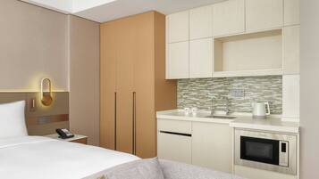 A modern hotel room with a bed, bedside table with a phone, a sofa, a kitchenette with a microwave, and a tiled backsplash.