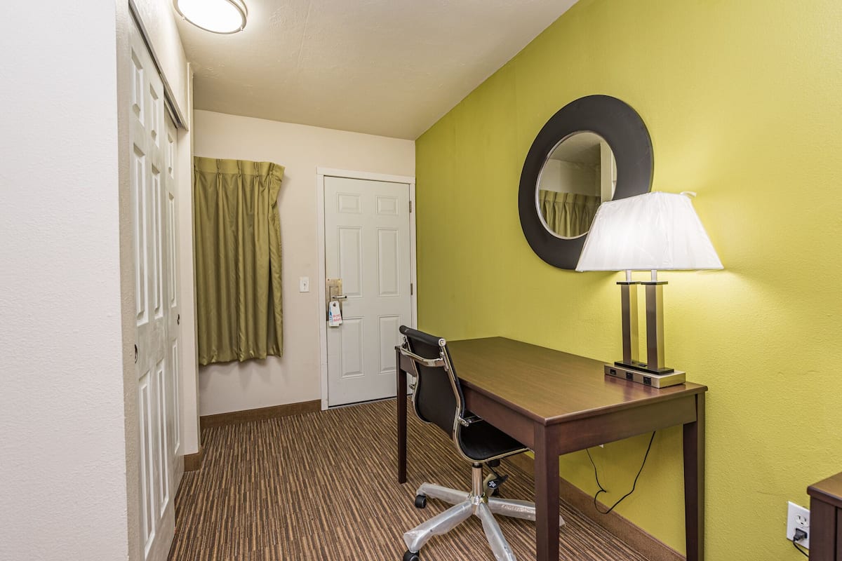 Habitación de hotel con un escritorio de madera, una silla, una lámpara, un espejo, una puerta y una cortina verde.