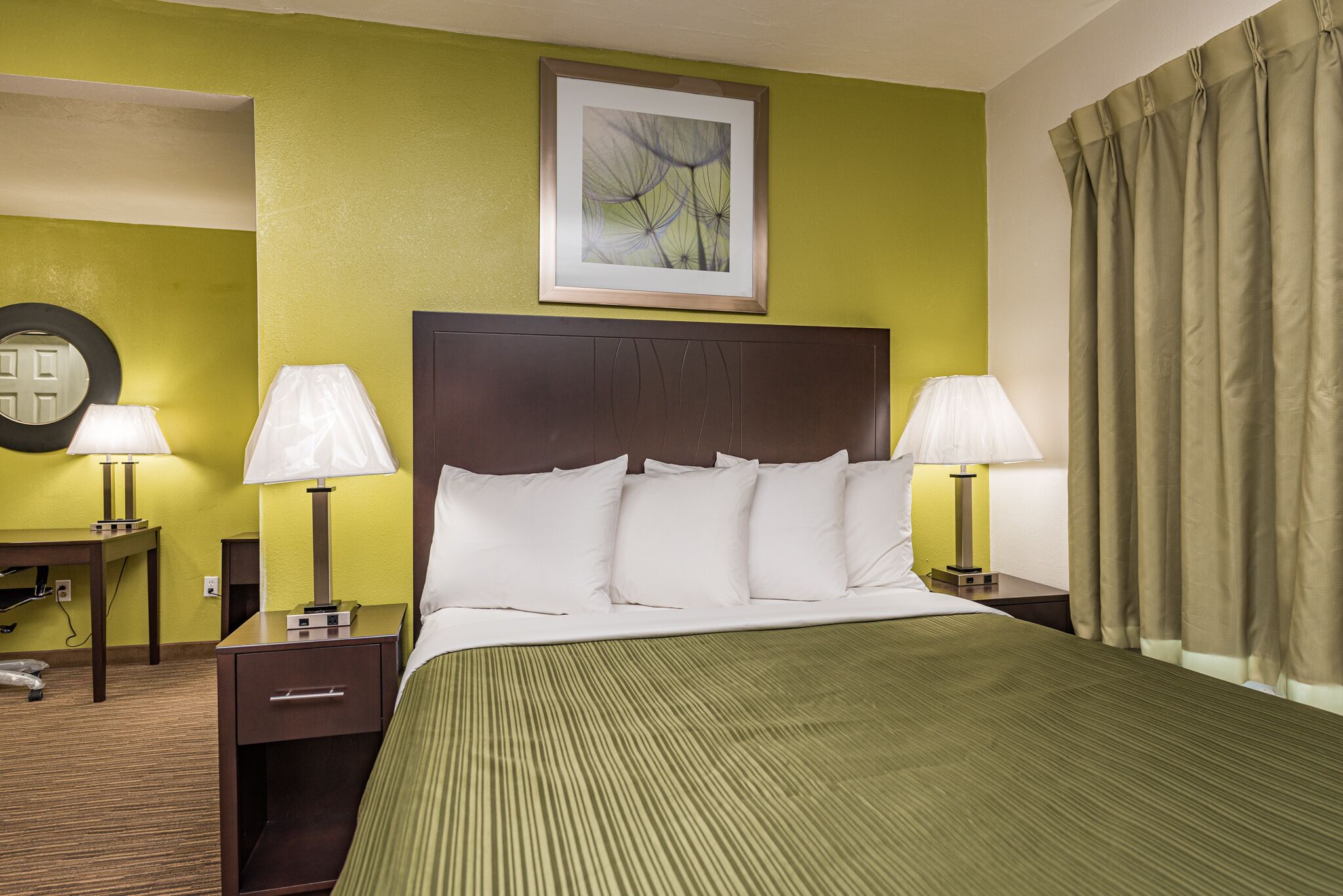 Habitación de hotel con una cama grande, dos mesitas de noche con lámparas, un cuadro enmarcado en la pared y una ventana con cortinas.