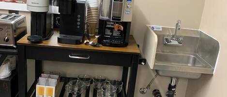 A kitchenette with a coffee machine, a sink, and a storage shelf with cups and a kettle.