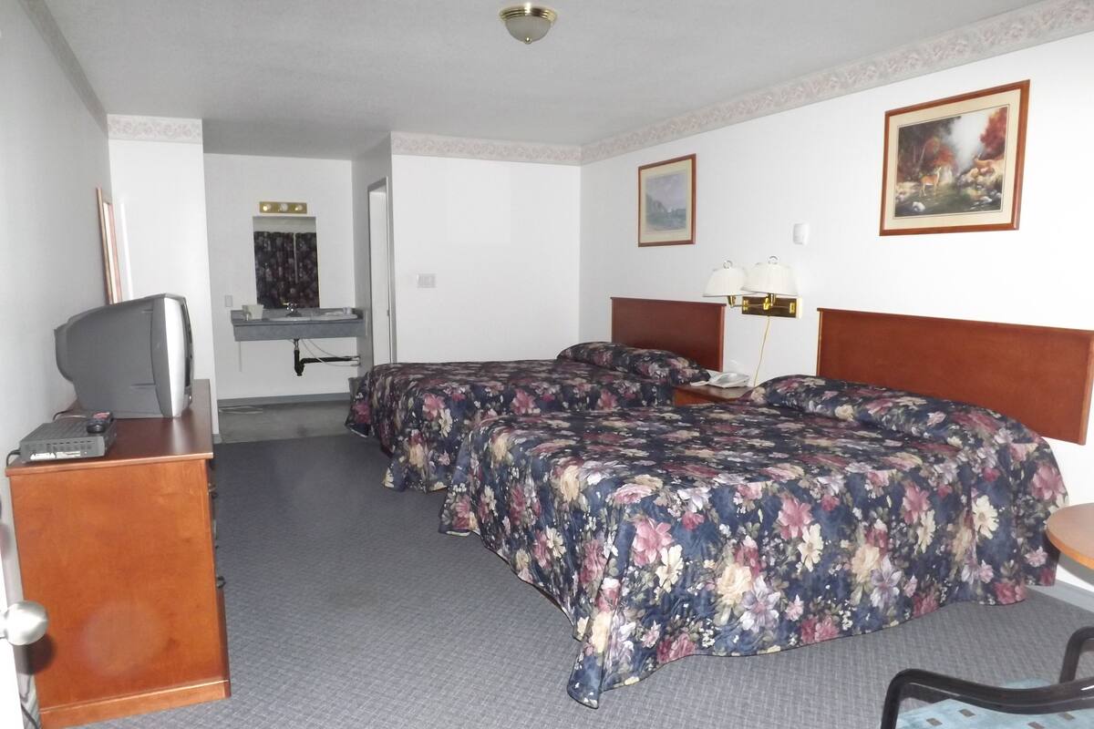 A hotel room with two beds, a desk with a computer, a chair, a TV, and a bathroom visible in the background.