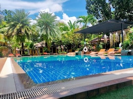 Una piscina con sillones de descanso, palmeras y una zona cubierta con plantas colgantes.