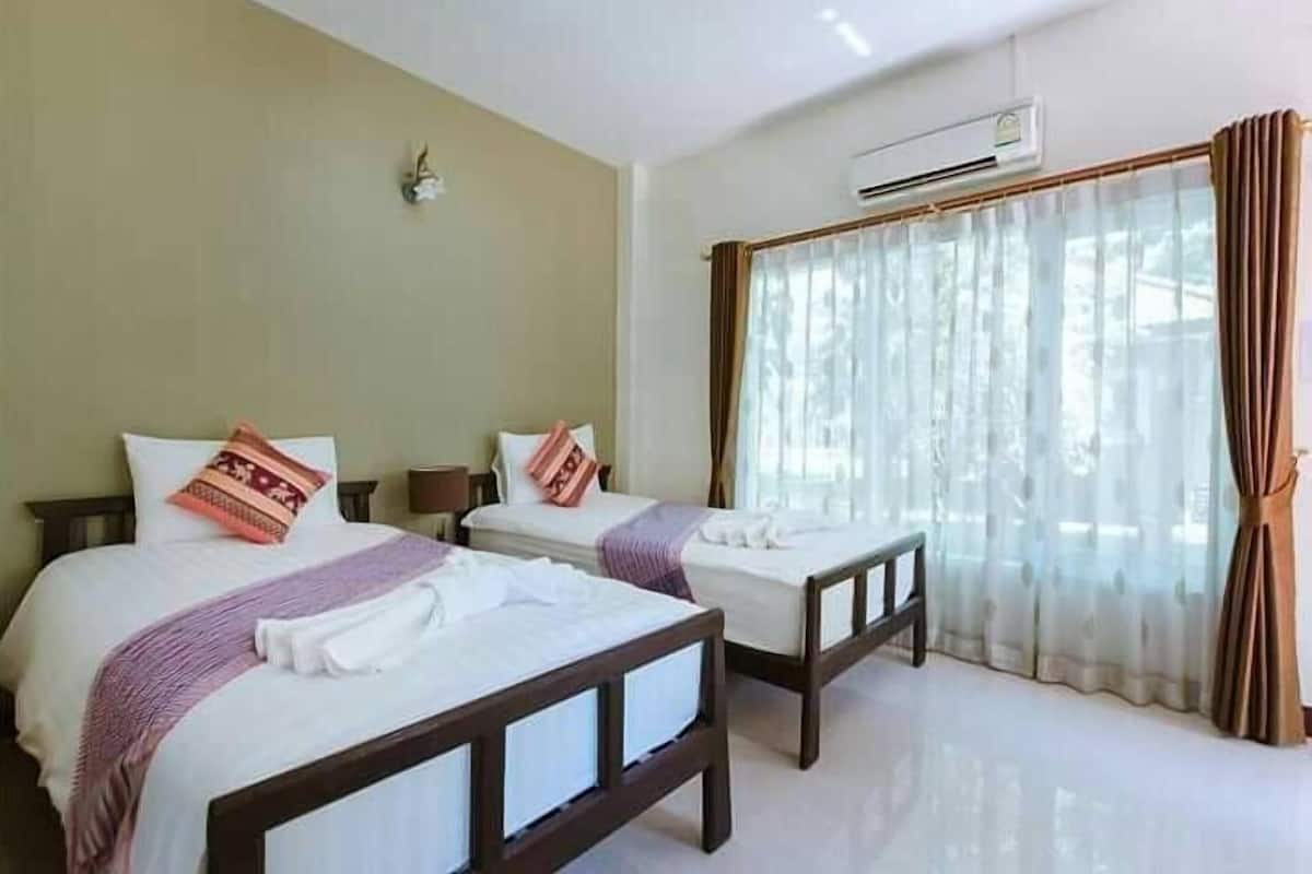 A hotel room with two beds, a window with curtains, an air conditioning unit, and a wall-mounted light fixture.