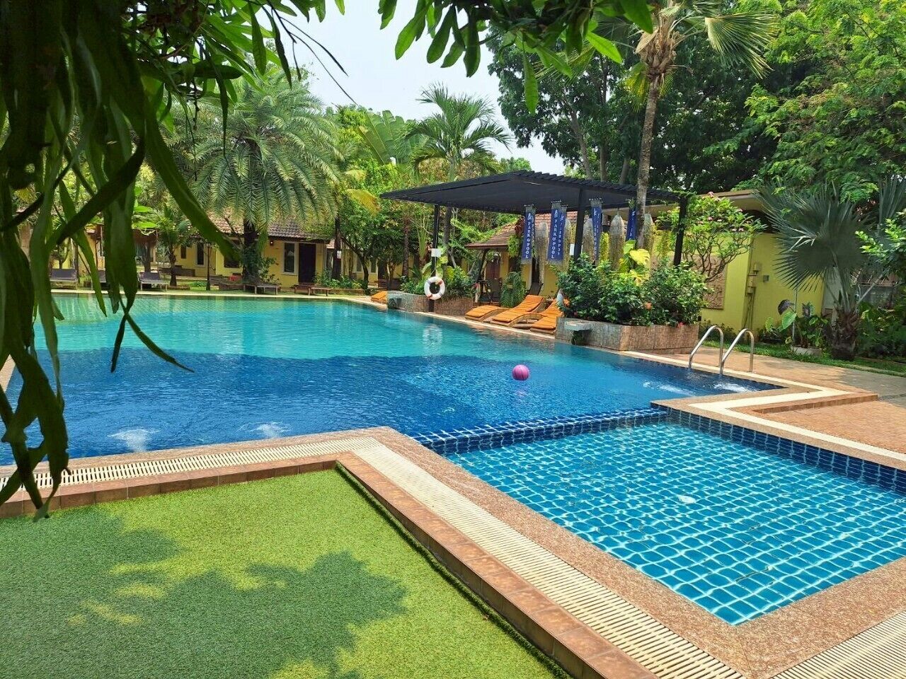 A swimming pool with a tiled deck, surrounded by greenery and a covered seating area.