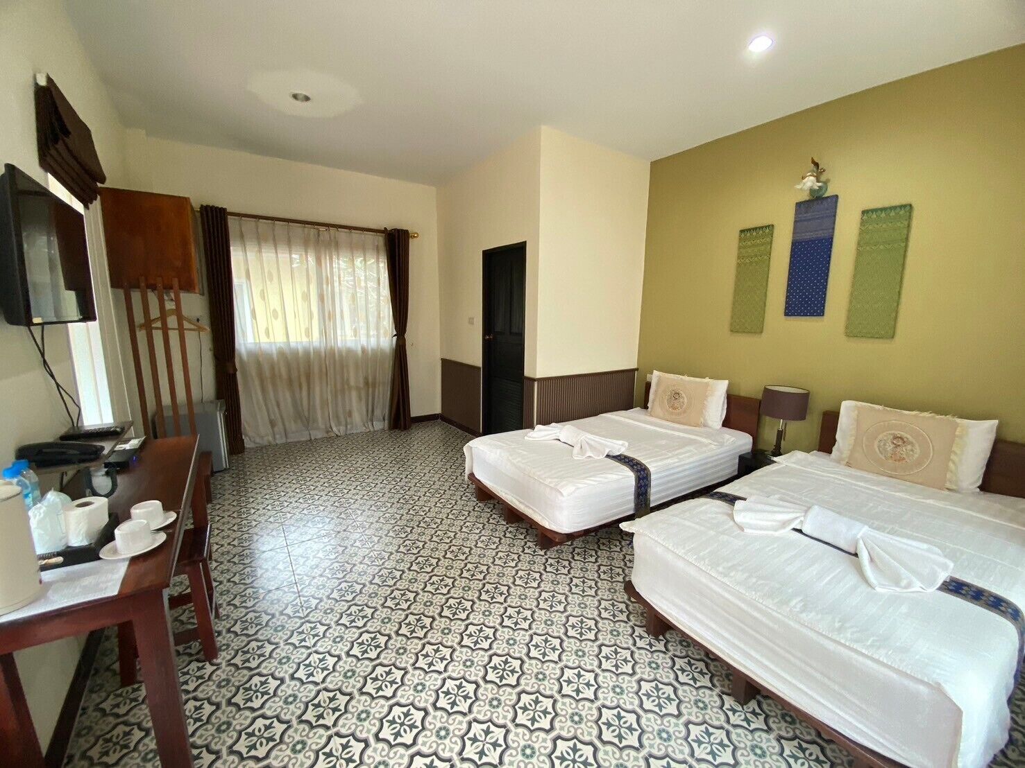 Habitación de hotel con dos camas, un escritorio con silla, un televisor y baldosas de suelo con diseño.