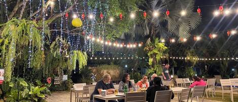 Un área de comedor al aire libre con mesas y sillas, rodeada de abundante vegetación y luces colgantes.
