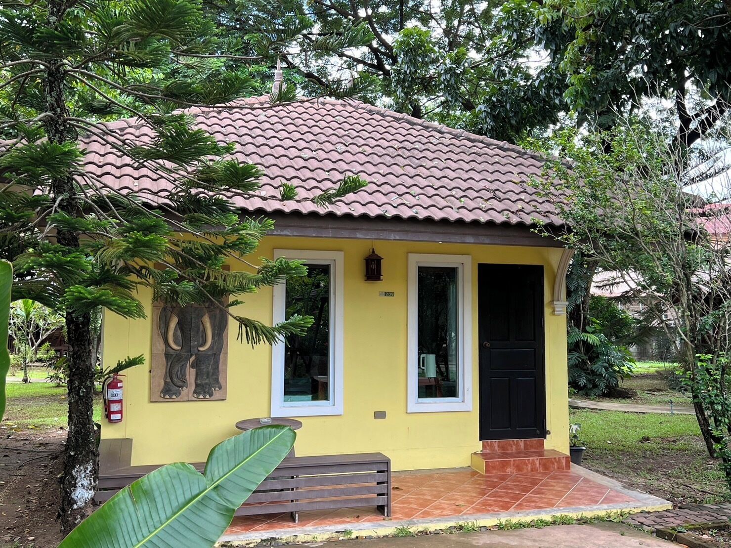Um pequeno edifício amarelo com telhado de azulejos, porta preta e um banco na frente.