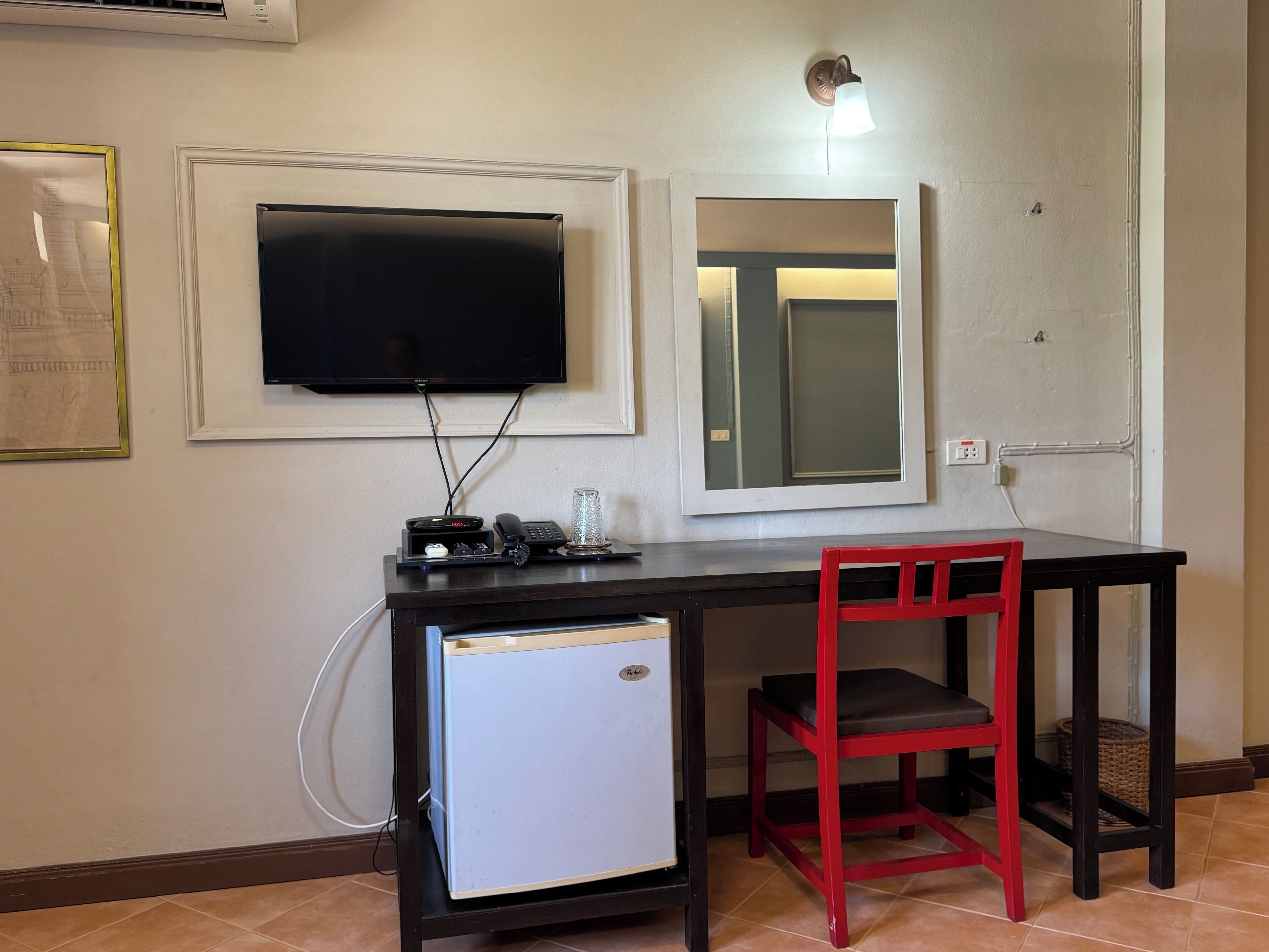 A compact room with a small desk, a television mounted on the wall, a mirror, a red chair, and a mini-fridge.