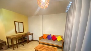 Quarto com escrivaninha de madeira, um banco, sofá com almofadas coloridas, luminária pendente e janela com cortinas.