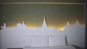 Uma cama com roupa de cama e almofadas brancas, uma parede lisa e um contorno de um horizonte urbano com uma estrutura imponente semelhante a um templo no centro.