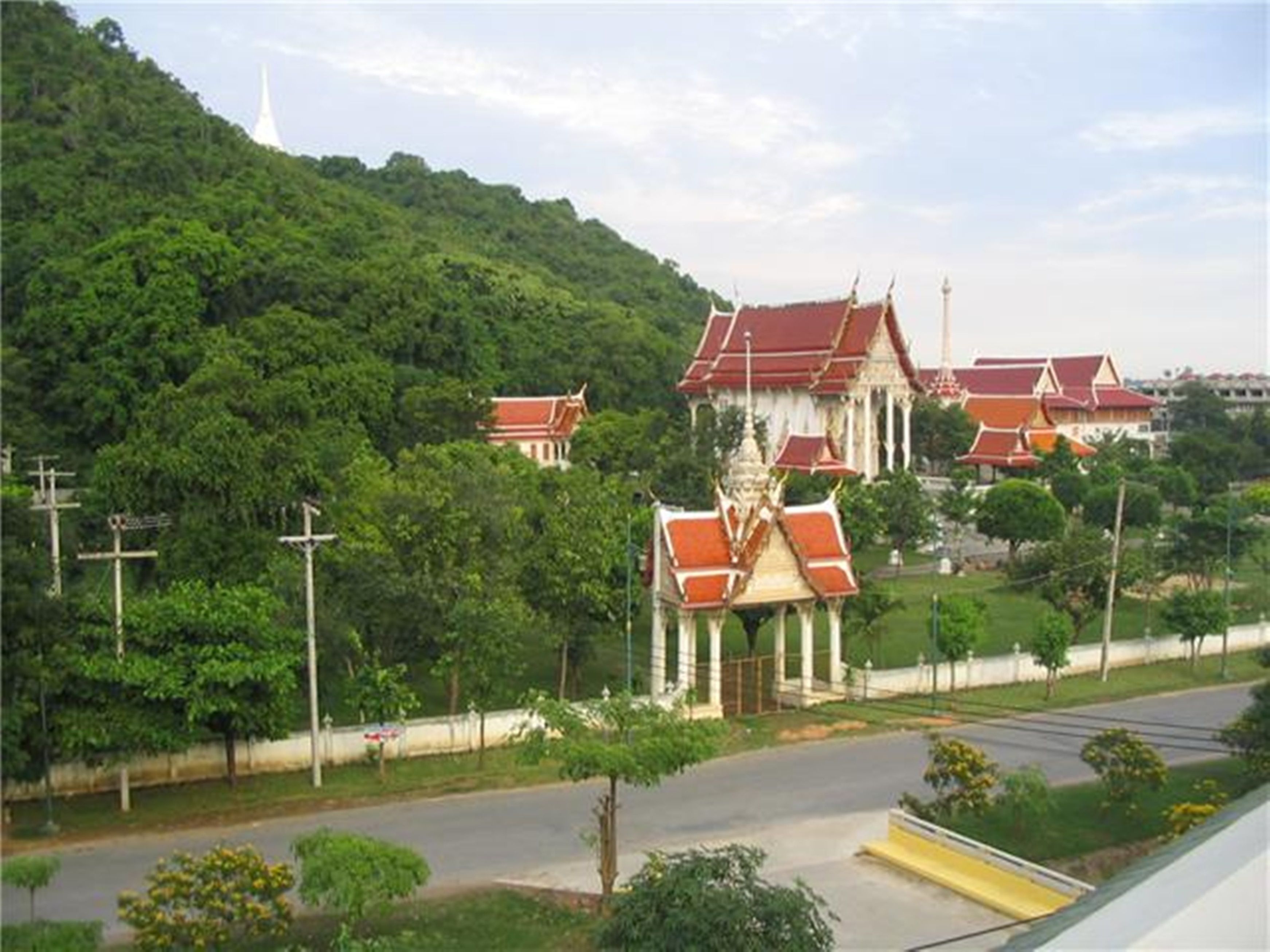 A traditional temple complex with multiple structures, surrounded by lush greenery and a road in the foreground.