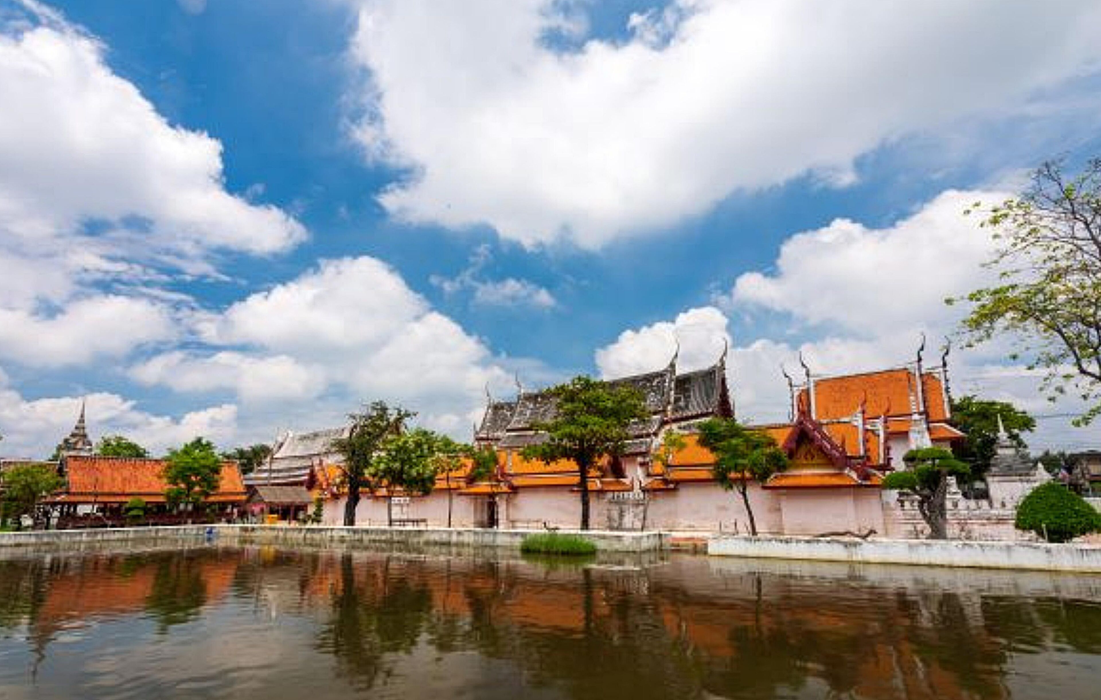 Un complejo de templos tradicional con varios edificios, una masa de agua tranquila al frente y un cielo despejado con nubes dispersas.