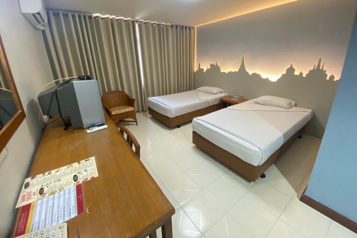 A hotel room with two beds, a desk with a computer, a chair, and a wall with a cityscape mural.