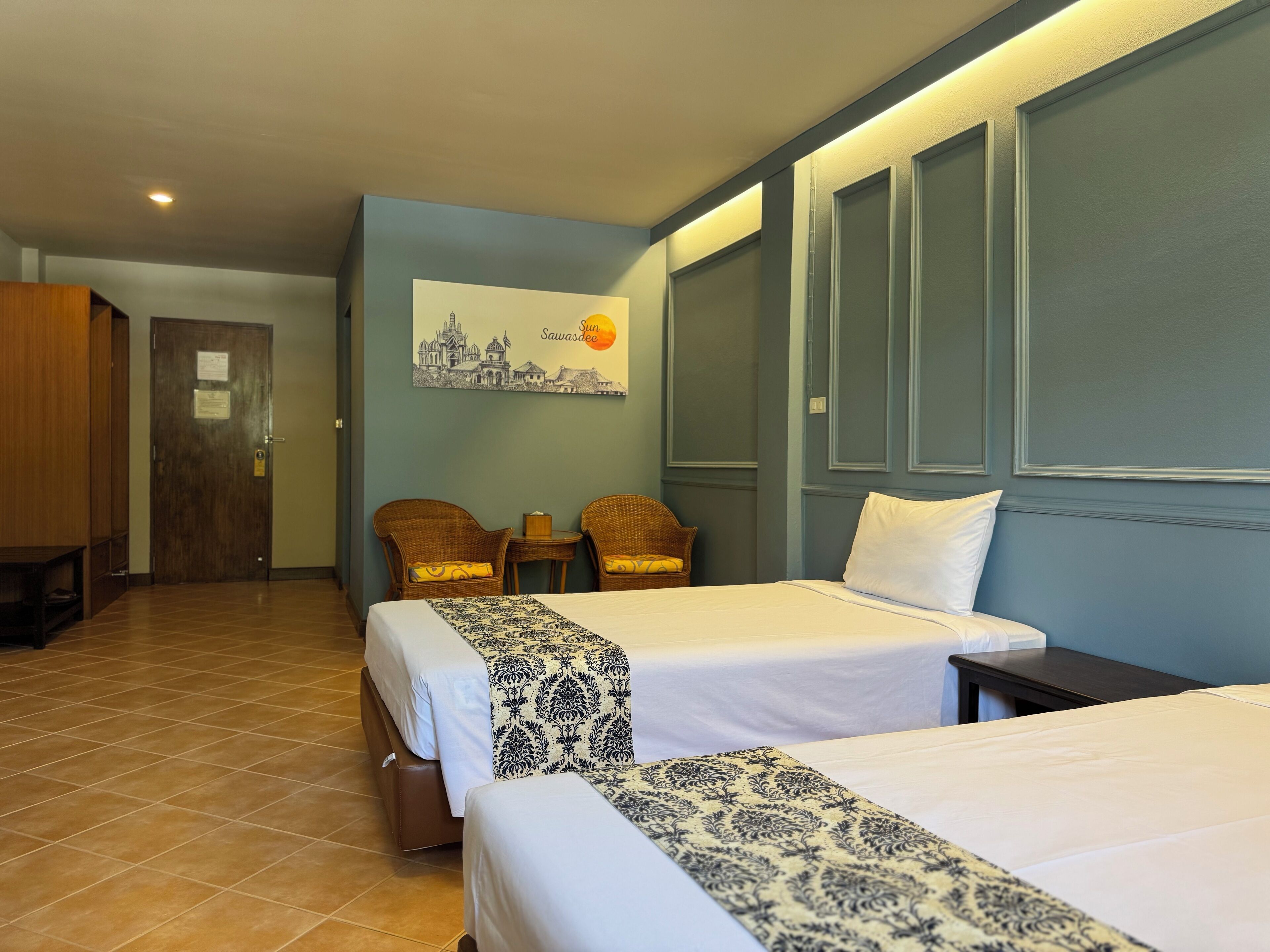 Una habitación de hotel con dos camas, un armario de madera, una mesita pequeña, dos sillas de mimbre y un cuadro colgado en la pared.