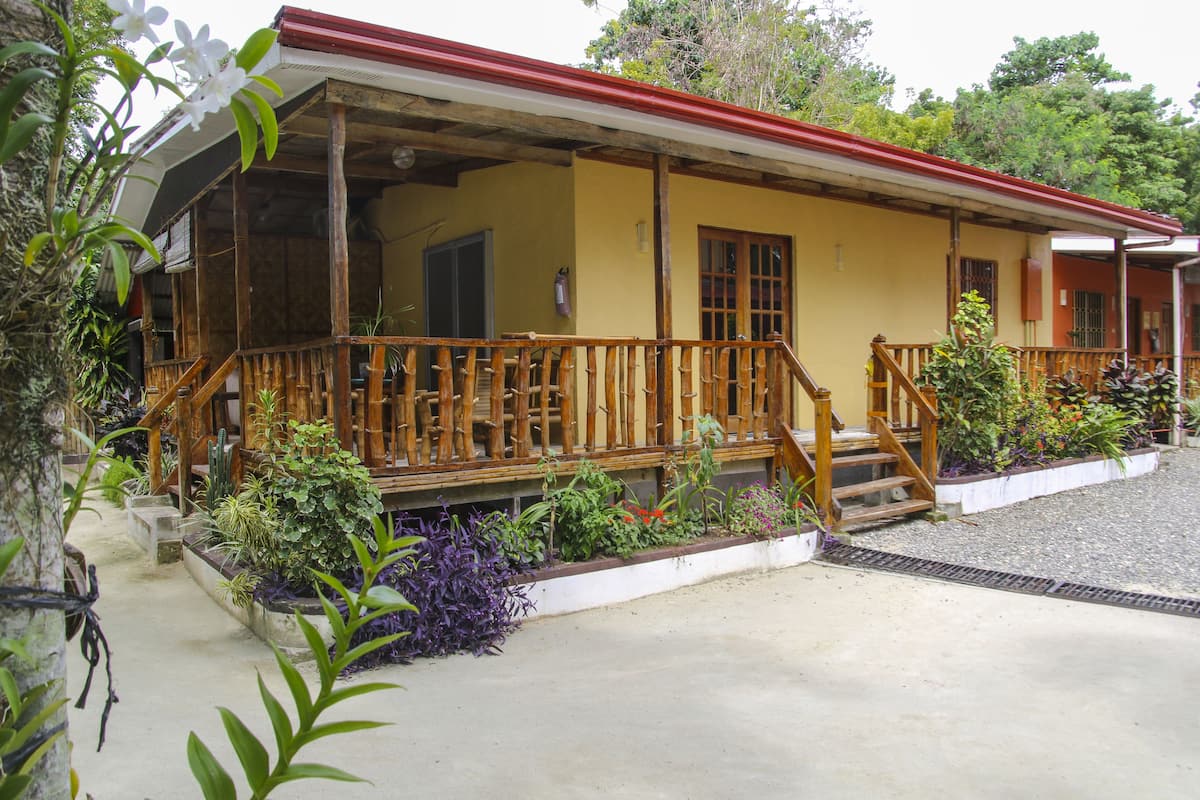 A yellow building with a red roof, wooden balcony, and surrounding plants.
