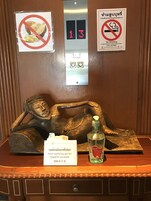 Uma estátua de madeira de uma figura reclinada, um dispensador de gel desinfetante para as mãos e um aviso de proibido fumar na parede.