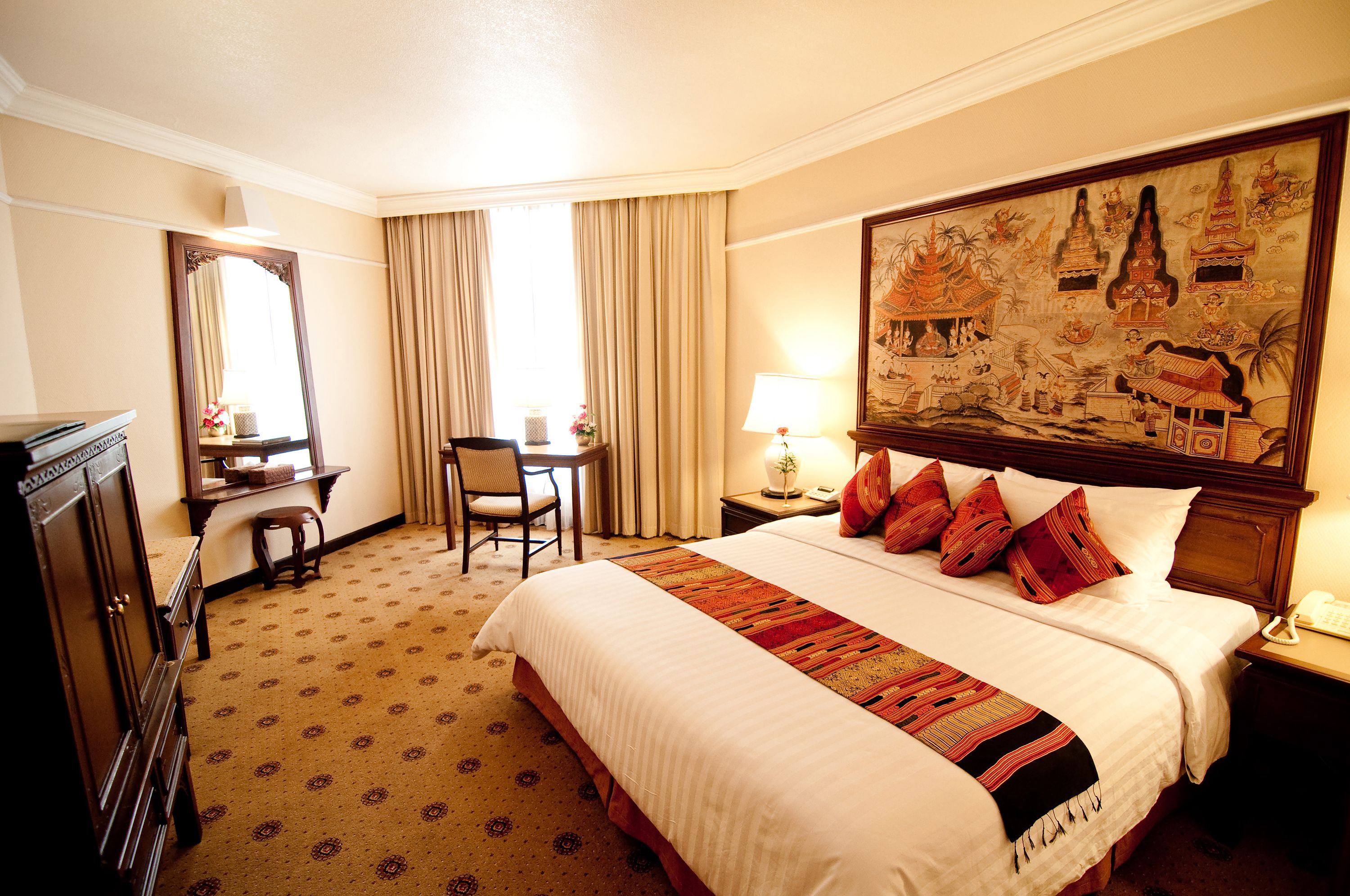 A hotel room with a large bed, a desk with a chair, a desk lamp, a telephone, and a painting on the wall.