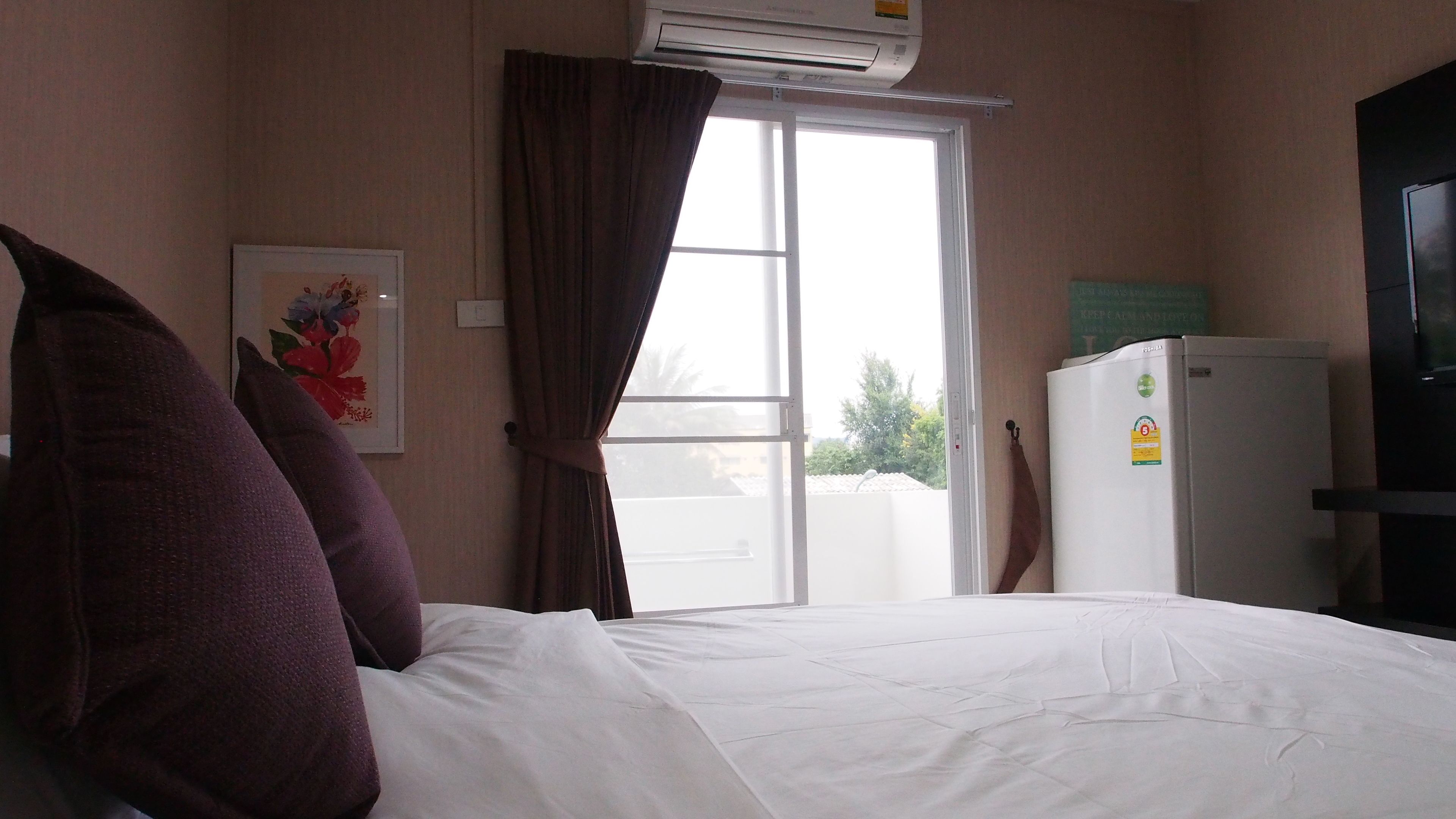 Une chambre d’hôtel comprenant un lit, une fenêtre avec des rideaux, un appareil de climatisation, un réfrigérateur et un téléviseur fixé au mur.