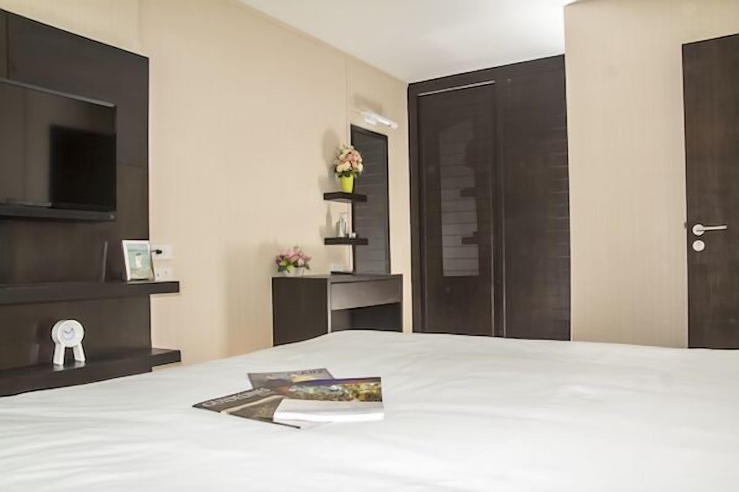 Una camera d'albergo con un letto, una televisione a muro, una scrivania con una sedia, uno scaffale con un vaso di fiori e una porta.