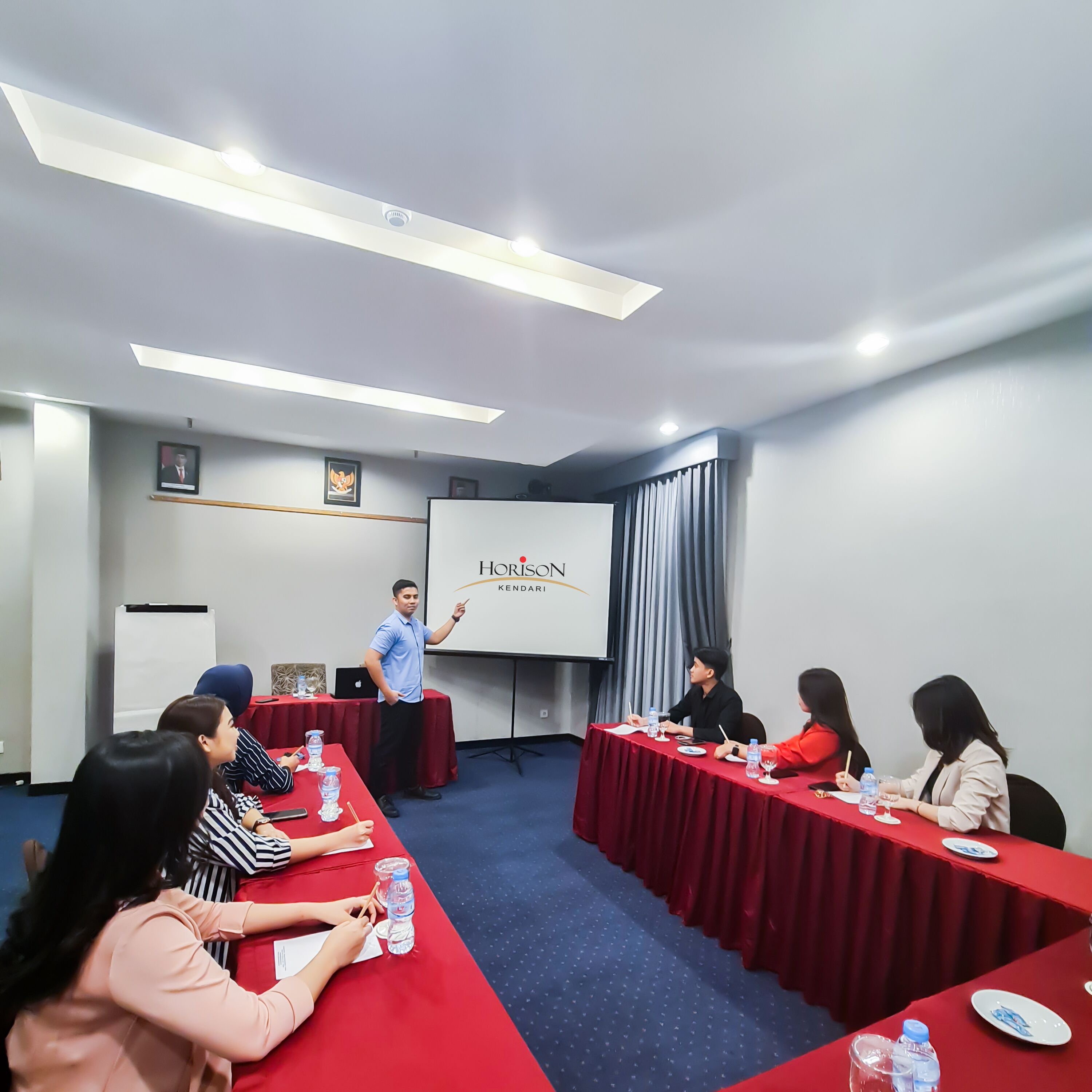 A conference room with a red table, attendees, and a presentation screen.