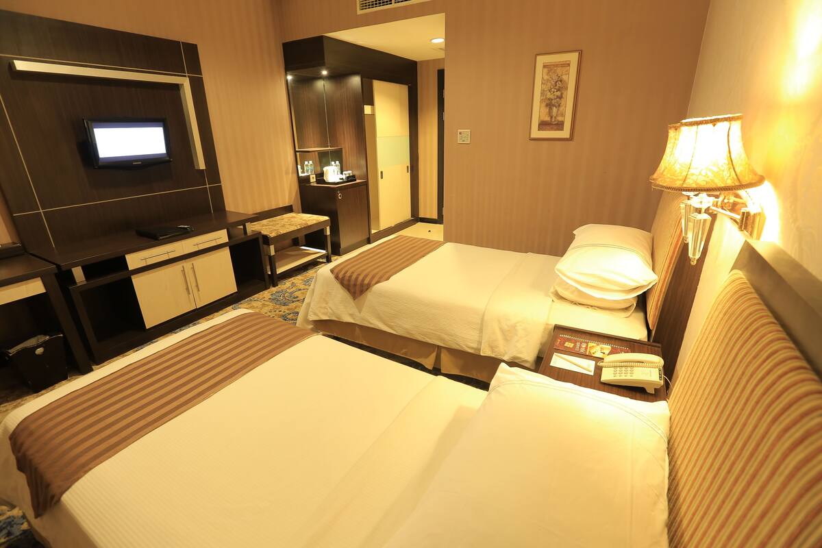 A hotel room with two beds, a desk with a flat-screen TV, a lamp, and a small table with a phone and notepad.