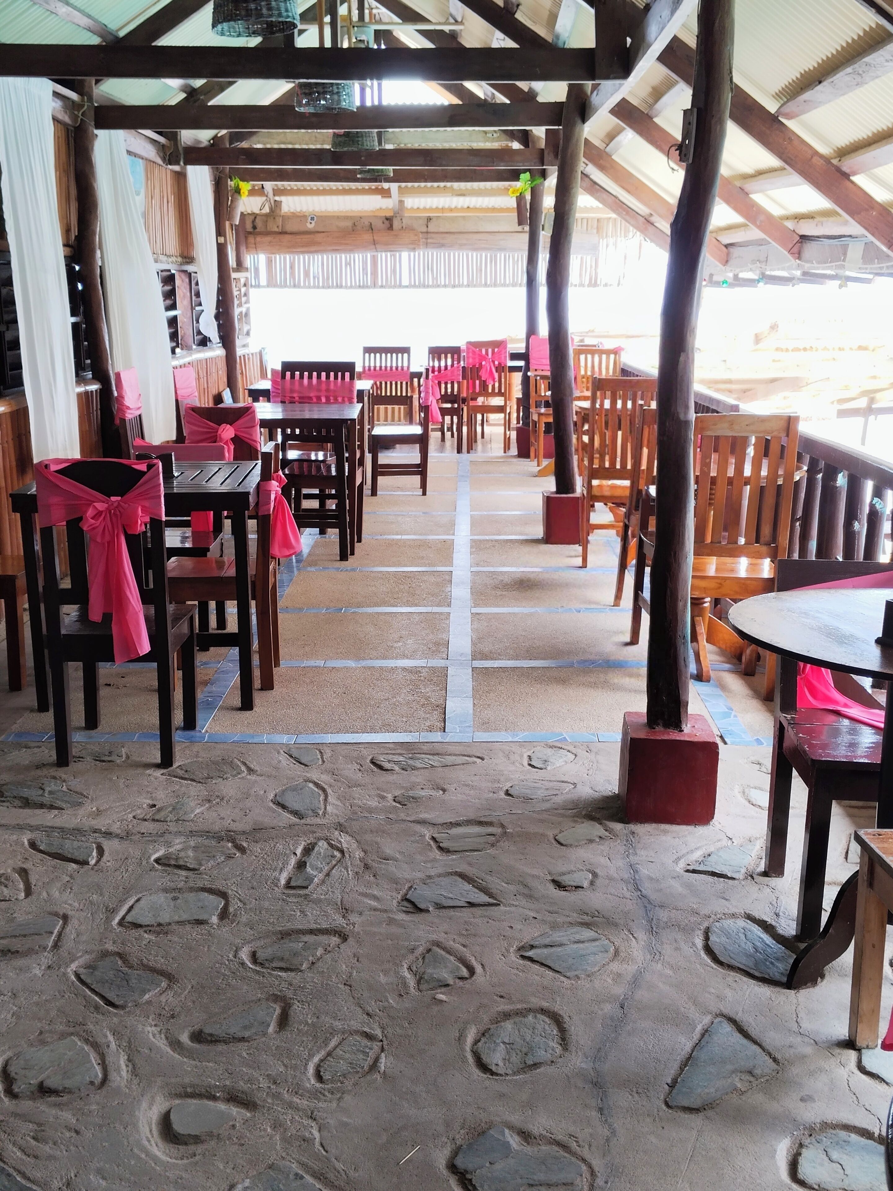A covered outdoor seating area with wooden tables and chairs, some with pink sashes, and a stone-patterned floor.