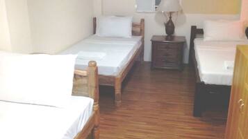 Quarto de hotel com duas camas de solteiro, piso de madeira, mesa de cabeceira pequena com abajur e ar condicionado na parede.