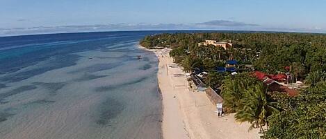 An aerial view of a beach with clear waters, a sandy shore, and buildings with red roofs.