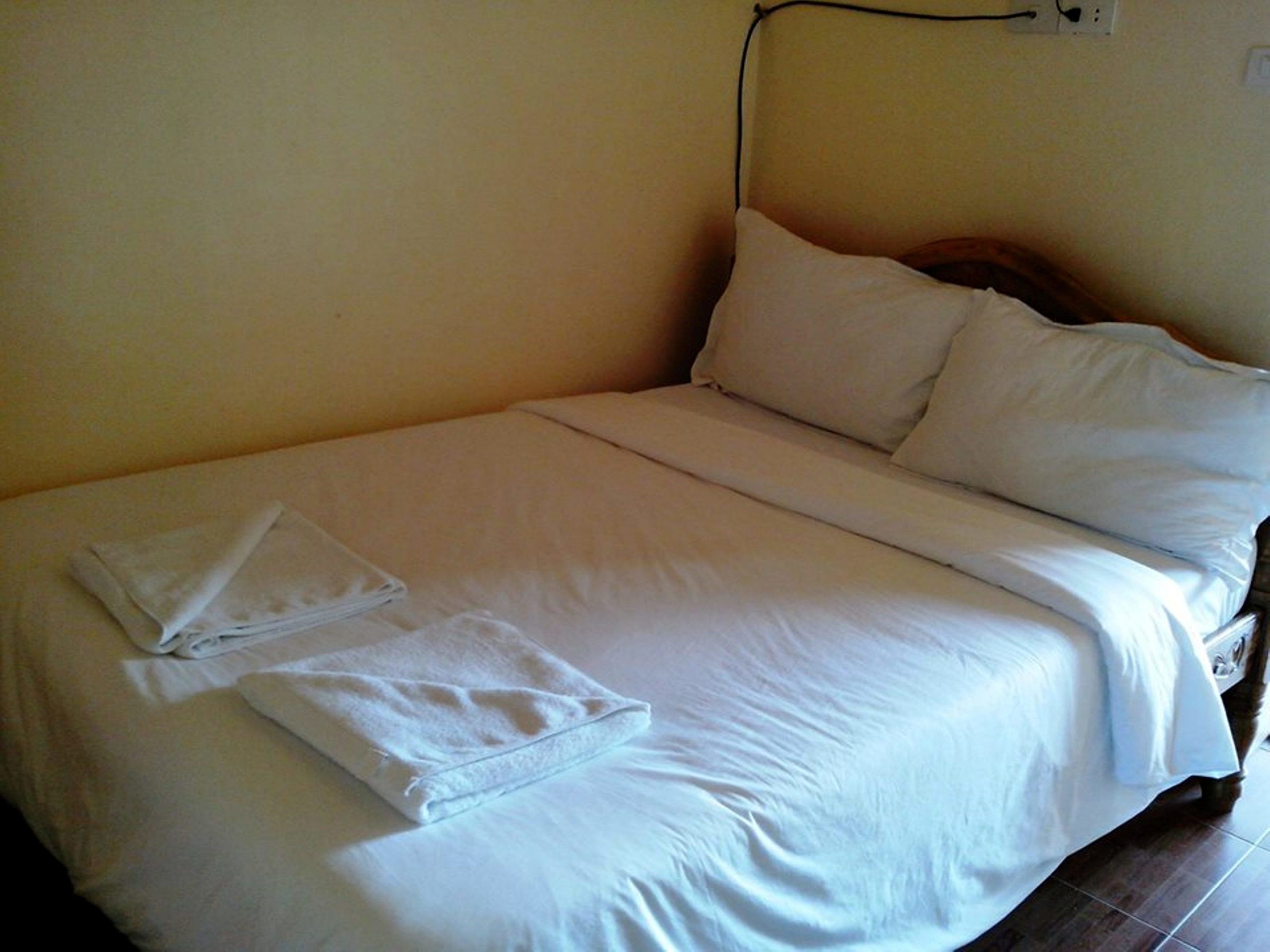 A neatly made bed with white linens, a folded towel, and a power outlet on the wall.