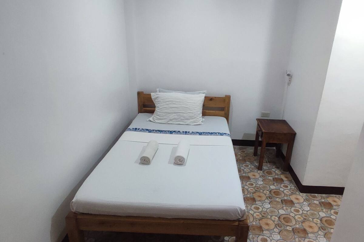 Una habitación pequeña y limpia con una cama individual, cabecera de madera, una mesita de noche y suelo estampado.