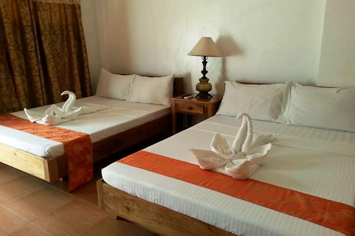 Dos camas dobles con sábanas blancas y detalles decorativos anaranjados, una mesita de noche de madera con lámpara, y una decoración de toalla con forma de cisne en cada cama.