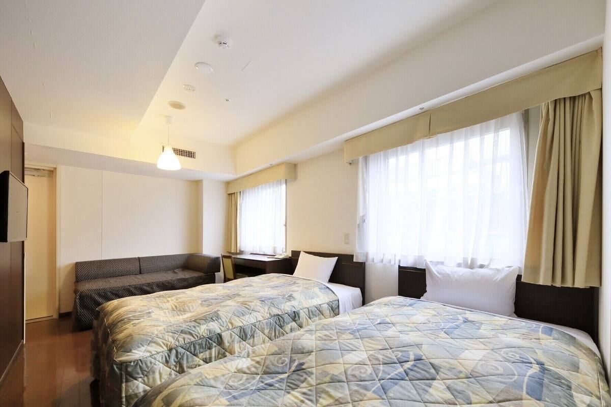Habitación de hotel con dos camas individuales, un sofá, un escritorio y un ventanal con cortinas.