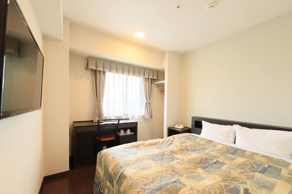 Habitación de hotel con cama, escritorio, silla, televisor y ventana con cortinas.