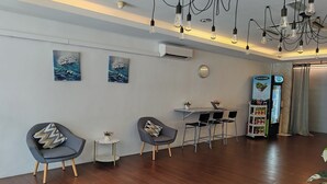 Un área de descanso moderna con suelo de madera, dos sillones grises con cojines estampados, una pequeña mesa redonda con una planta, una unidad de aire acondicionado blanca y una máquina expendedora.