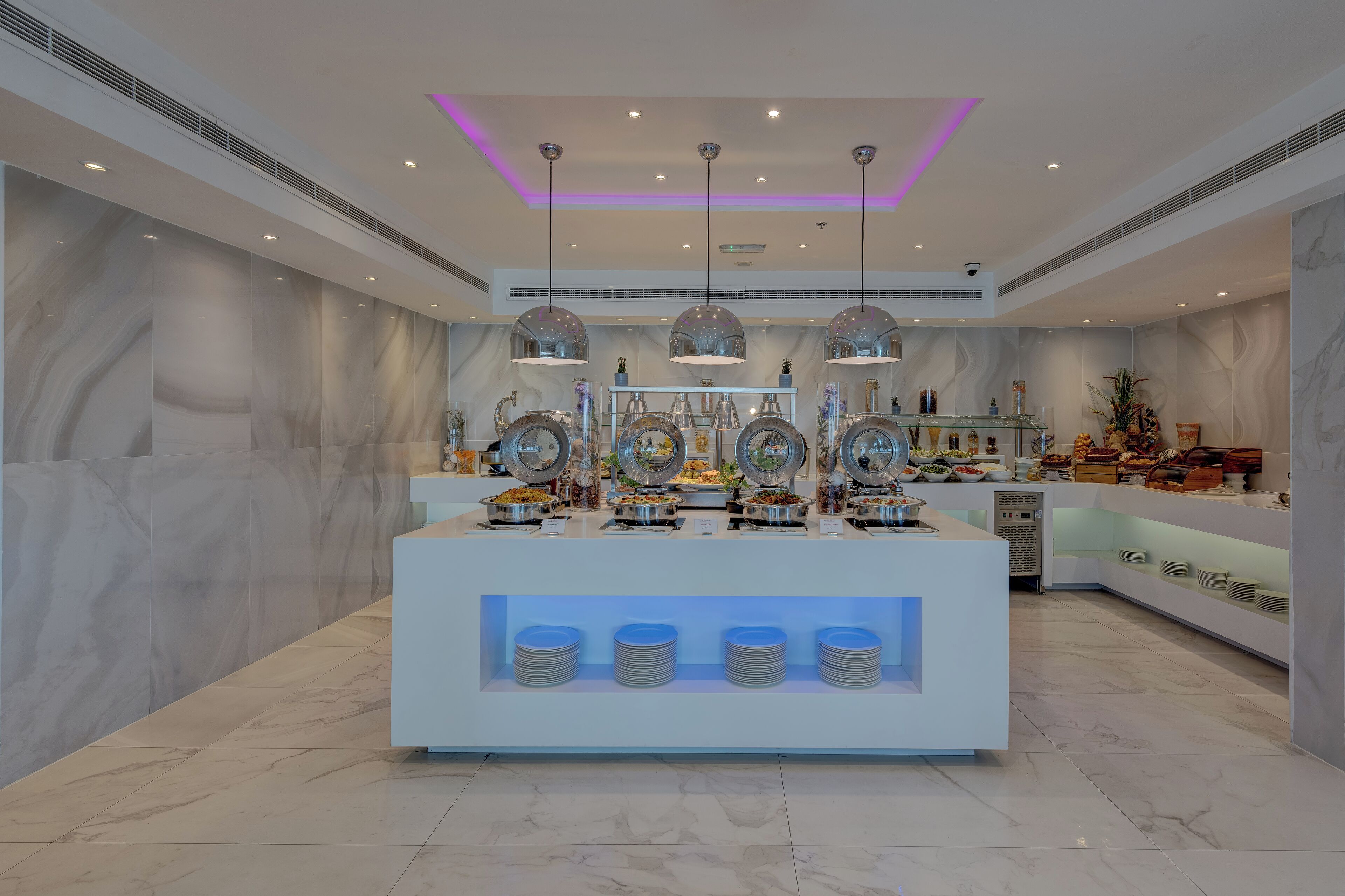 Un área de buffet moderna con paredes de mármol, iluminación colgante y estaciones de comida ordenadas.
