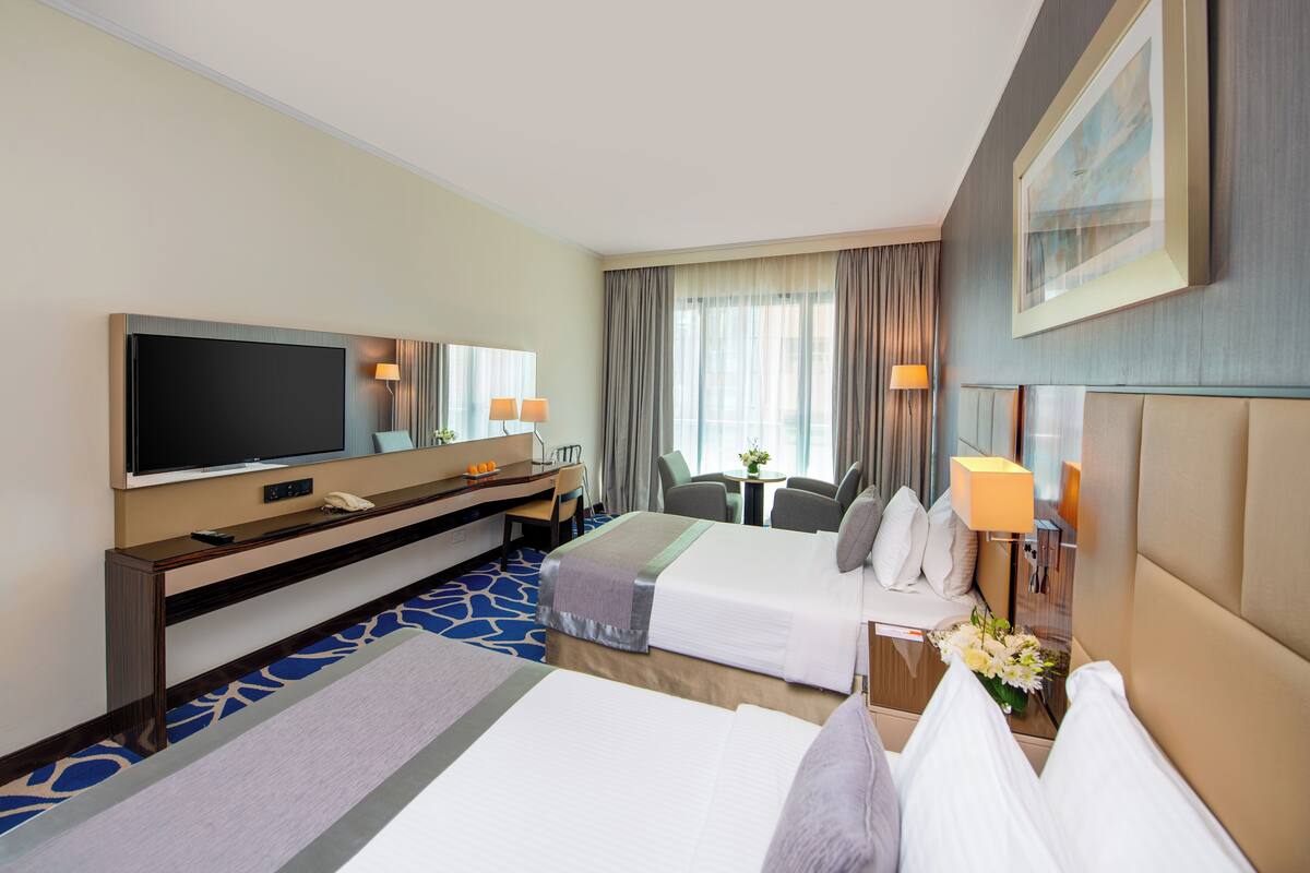 Une chambre d’hôtel avec deux lits, une télévision, un bureau avec une chaise, une petite table avec une lampe et une fenêtre avec des rideaux.
