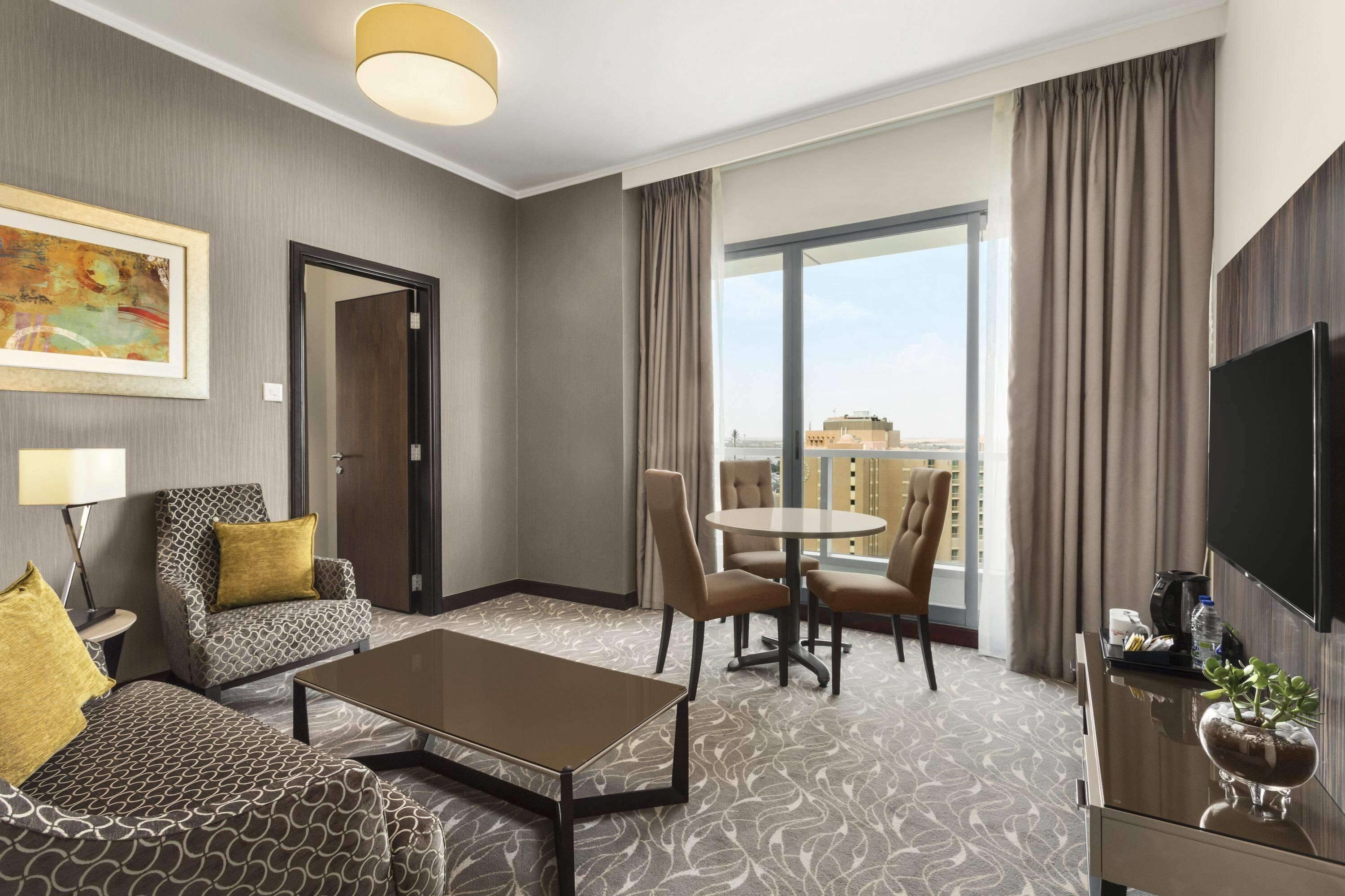 Habitación de hotel con sofá, mesa de centro, mesa de comedor con sillas, televisión y balcón con vistas.