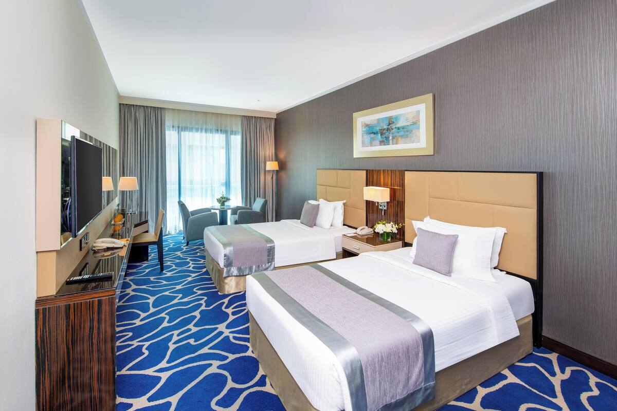 Une chambre d’hôtel avec deux lits, un bureau avec un ordinateur, une chaise, une télévision et une grande fenêtre avec des rideaux.