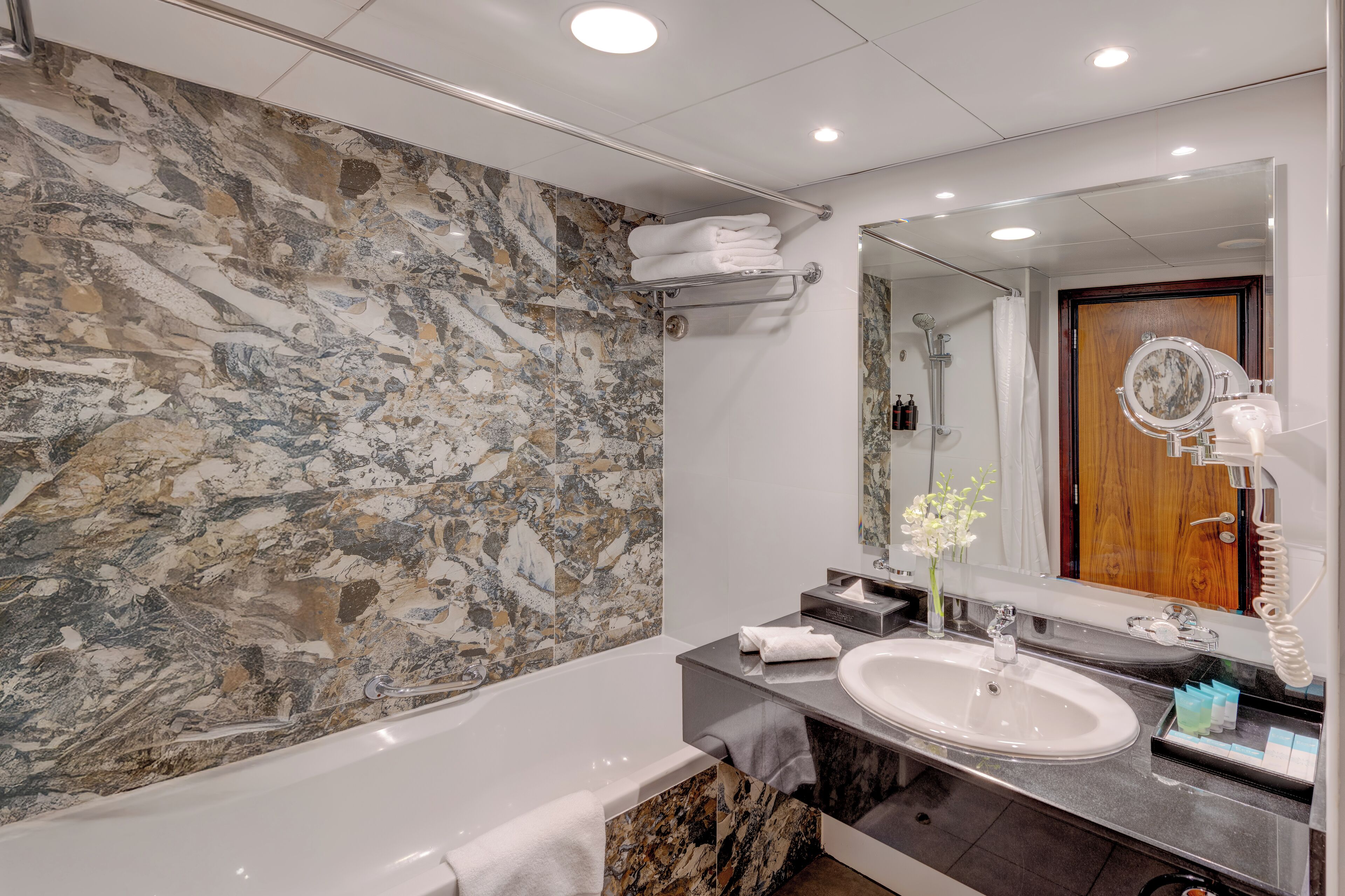 A modern bathroom with a marble wall, a bathtub, a sink, and a vanity mirror.