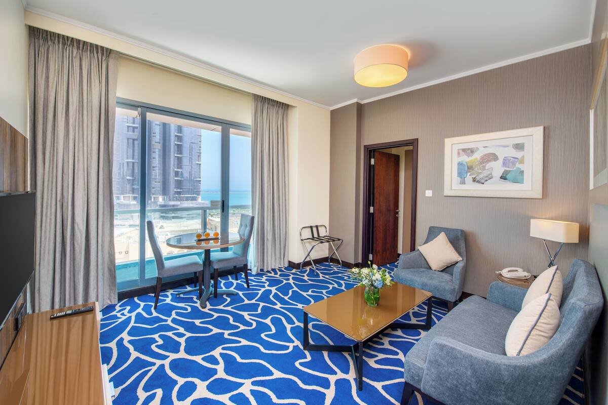 Une chambre d’hôtel avec une grande fenêtre, une moquette bleue à motifs, un canapé, une table basse et un téléviseur.