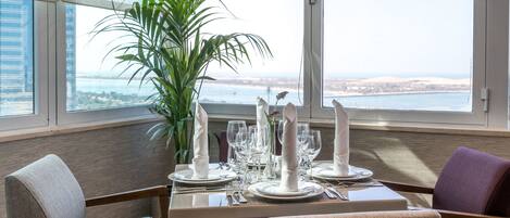 Een eettafel gedekt voor vier personen, met wit servies, een potplant en uitzicht op de stad.