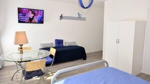 Une chambre d’hôtel moderne avec une table à manger en verre, un lit recouvert d’une couverture bleue, un téléviseur fixé au mur et une armoire.