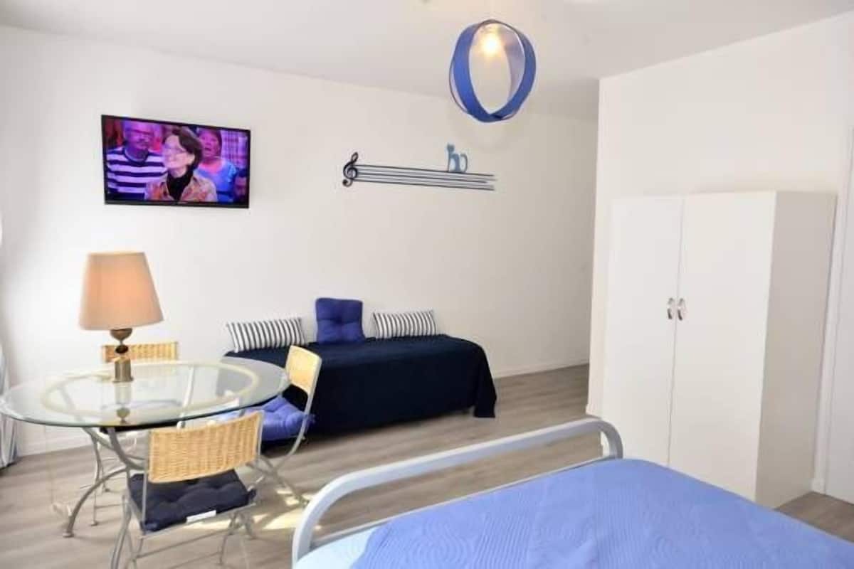 A modern hotel room with a glass dining table, a bed with a blue bedspread, a television mounted on the wall, and a wardrobe.