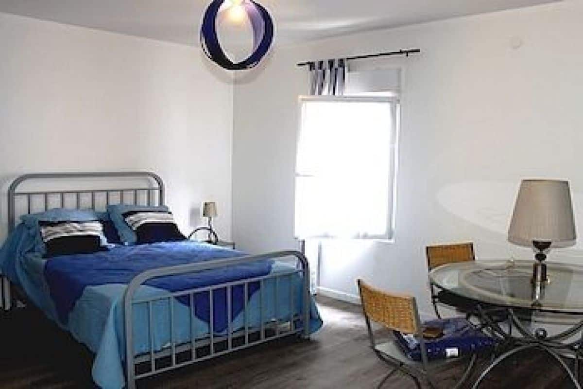 A bedroom with a metal bed frame, a round glass table with a lamp, and a window with curtains.