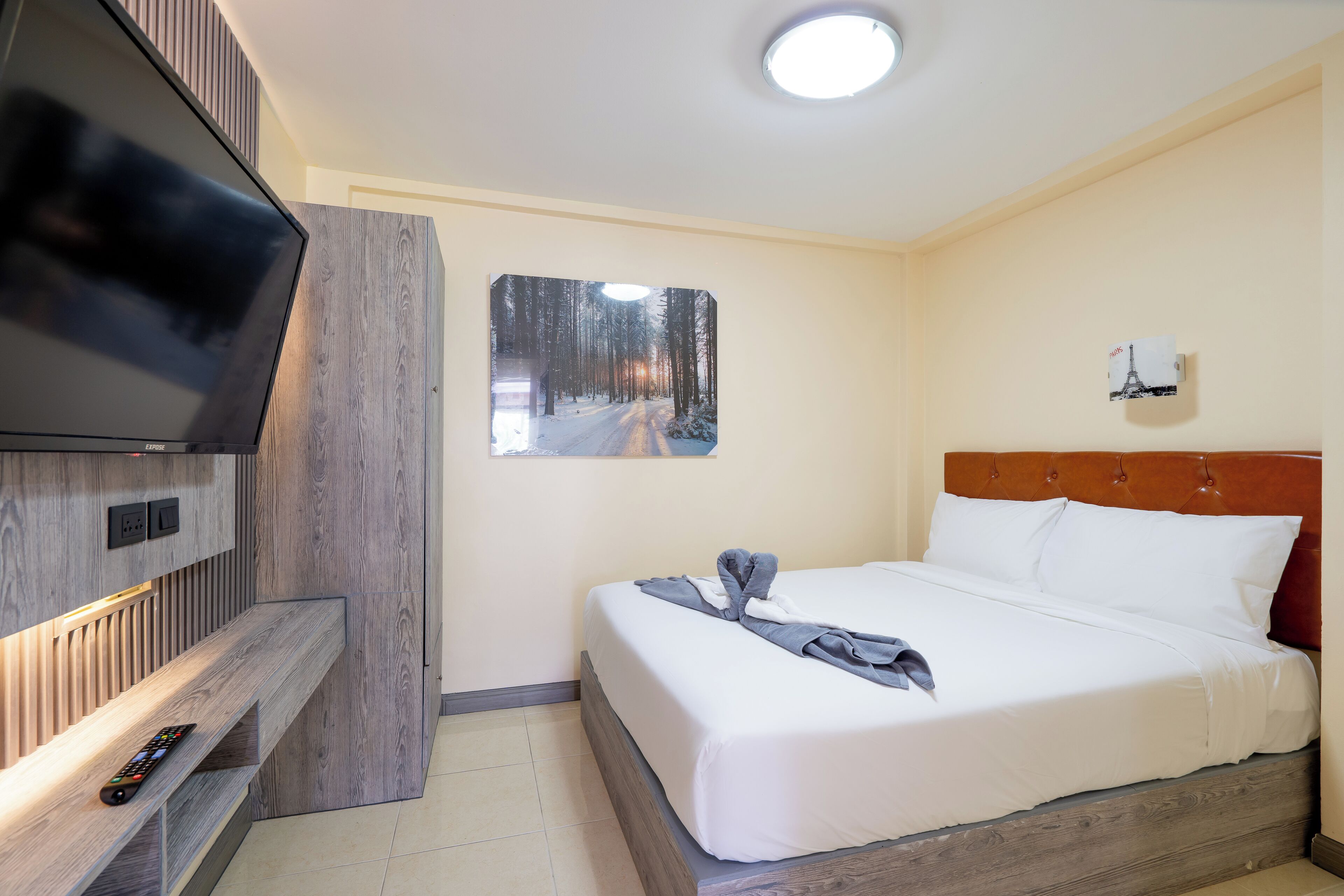 A hotel room with a bed, a TV mounted on the wall, a painting of a snowy forest, and a wooden headboard.